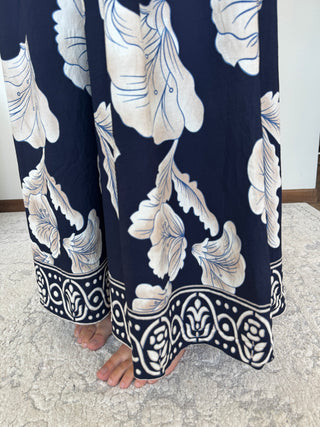 A close-up of a person's bare feet wearing navy blue wide-leg pants with a large cream floral print and a decorative border hem, standing on a textured light gray rug.