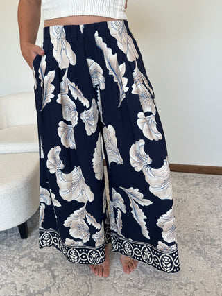 A person wearing wide-leg navy pants with a large white and light blue floral print and a decorative border at the hem, standing indoors on a patterned rug.
