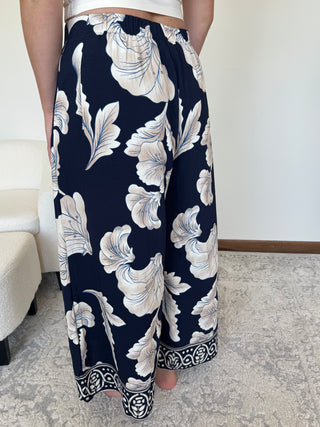 A person wearing wide-leg navy pants with a large white floral print and a decorative border at the hem, standing indoors on a patterned rug.