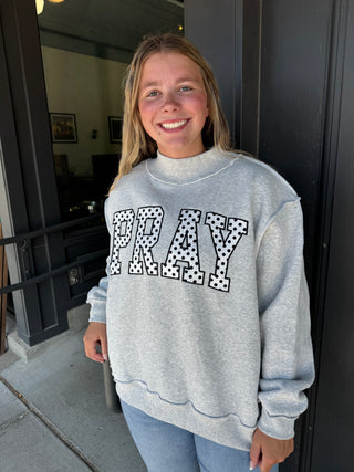 A smiling woman wearing a light gray crewneck sweatshirt with the word 'PRAY' in large black-and-white polka dot outlined letters, standing outdoors in front of a dark doorway.
