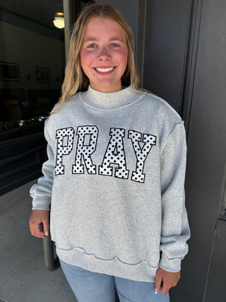 A smiling young woman wearing a light gray mock-neck sweatshirt with 'PRAY' in black-and-white polka dot block letters, paired with light blue jeans, standing in front of a dark doorway.