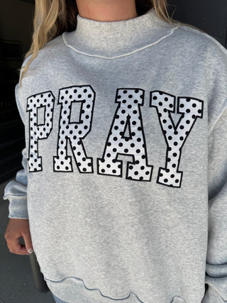 A close-up of a person wearing a light heather gray mock-neck sweatshirt with the word 'PRAY' in large varsity-style letters across the chest. The letters are white with black polka dots and black outlines.