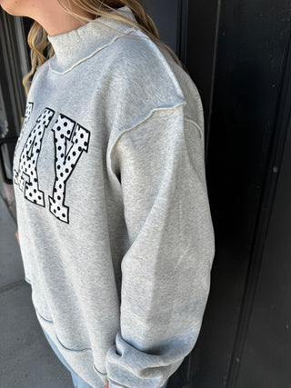 A person wearing a light heather gray sweatshirt with large black-and-white polka dot block letters across the chest, shown in a close-up outdoor shot against a dark door.