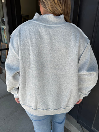 A person viewed from behind wearing an oversized light heather gray sweatshirt with a mock neck and ribbed hem, paired with light blue jeans, standing in front of a dark doorway.