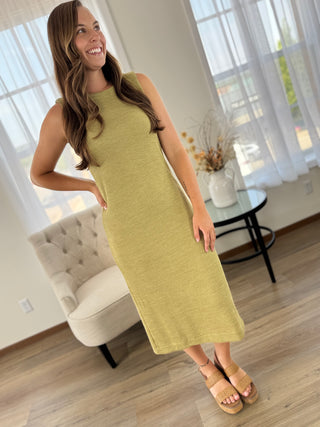 A woman modeling a sleeveless, high-neck, knee-length olive green dress in a bright, modern living room setting.