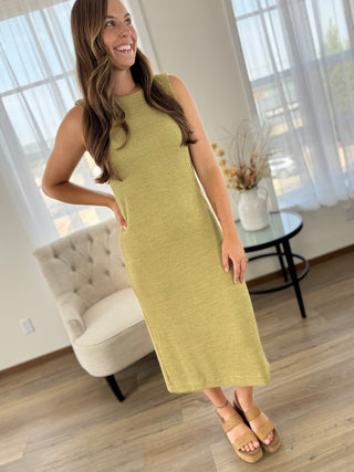 A woman modeling a sleeveless, knee-length olive green knit dress in a bright, modern room with light wood floors, a cream tufted armchair, and sheer white curtains.