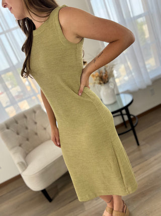A woman wearing a sleeveless olive green knit midi dress with side pockets, standing in a bright living room with a tufted armchair and a small table in the background.