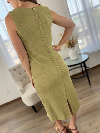 A woman wearing a sleeveless olive green midi dress with a back button closure, photographed in a bright indoor room with a chair, table, and window curtains in the background.