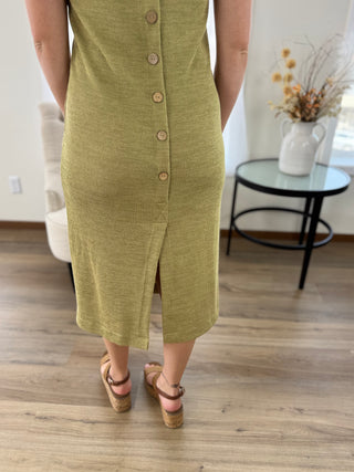 A person wearing a sleeveless olive green midi dress with a button-front closure and side slit, standing on a wood floor in a bright indoor room.