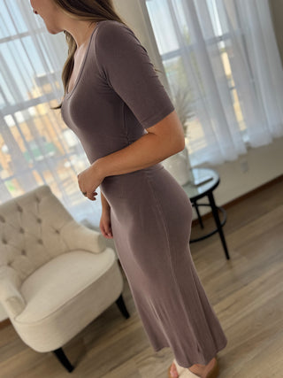 A woman wearing a fitted short-sleeve midi dress in a muted taupe-brown color, standing in a bright room with a tufted armchair and window curtains.