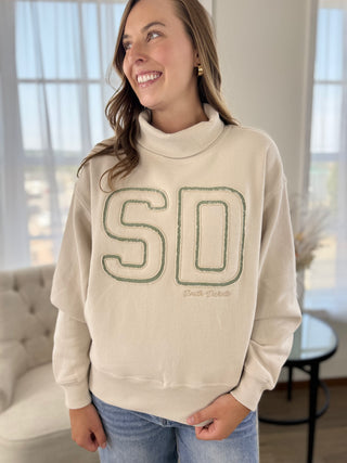 A woman wearing a cream-colored mock neck sweatshirt with 'SD' outlined in green across the chest, paired with blue jeans, standing in a brightly lit room.