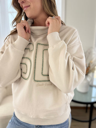 A woman wearing a cream-colored sweatshirt with a green graphic print of the number 50 and the words 'South Dakota' on the front, paired with blue jeans in a bright indoor setting.