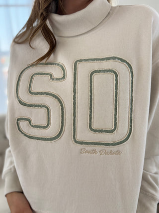 A close-up of a person wearing a cream-colored turtleneck sweater with a large embroidered 'SD' logo outlined in green and the words 'South Dakota' embroidered below.