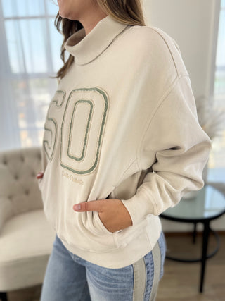 A woman wearing a cream-colored turtleneck sweatshirt with a large green outlined '50' graphic on the front, paired with light blue jeans, in a bright indoor setting.