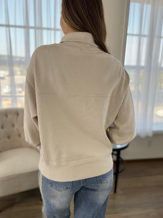 A woman viewed from behind wearing a light beige long-sleeve collared sweatshirt and blue jeans in a bright room with large windows and sheer curtains.