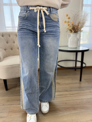 A person wearing light blue wide-leg jeans with beige side panels and a cream drawstring waist, standing in a bright indoor room with wood flooring, a tufted chair, and a small table with dried flowers.