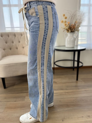 A person wearing light blue wide-leg jeans with two vertical beige side panels and a cream drawstring waist, standing in a bright indoor room with wood flooring, a tufted chair, and a small table with dried flowers.