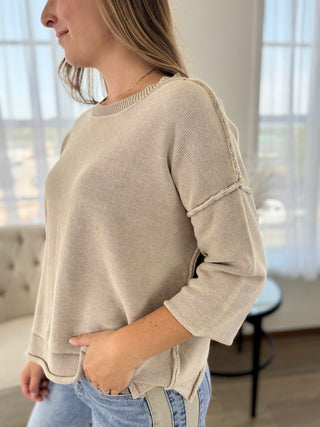 A woman wearing a light beige knit sweater with three-quarter sleeves and ribbed cuffs, paired with blue jeans in a bright, modern room.