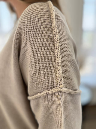 Close-up of a person wearing a beige knit sweater, showing the shoulder seam and sleeve construction with soft, natural lighting.