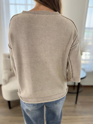 A woman viewed from behind wearing a light beige knit sweater and blue jeans in a bright indoor room with a chair and window in the background.