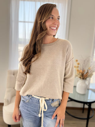 A woman wearing a light beige crewneck knit sweater and blue jeans with a white drawstring, standing in a bright, modern living room.