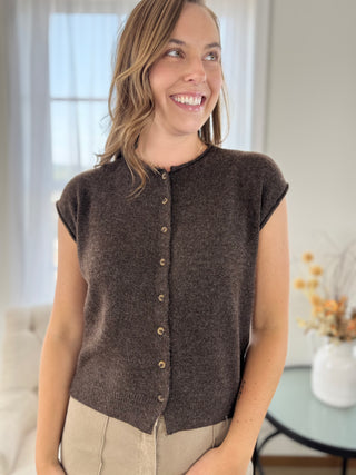 A woman wearing a dark brown sleeveless button-front knit top with a round neckline, shown in a bright indoor setting.
