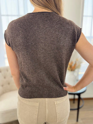 A woman viewed from the back wearing a short-sleeve knit top in a dark brown heather color, paired with light beige pants, in a bright indoor setting.