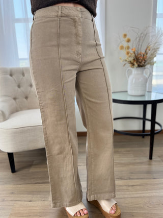 A person wearing beige wide-leg cropped pants with a dark brown top and beige wedge sandals, standing in a bright living room with a tufted chair and side table in the background.