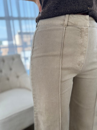 Close-up of a person wearing light beige straight-leg or bootcut pants with a dark charcoal knit top, standing in a bright room with a window and a tufted chair in the background.