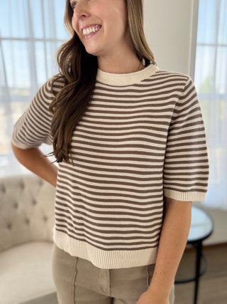 A woman wearing a brown and cream striped short-sleeve knit sweater, paired with beige pants, standing in a brightly lit room with a tufted sofa in the background.