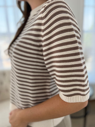 A close-up side view of a person wearing a short-sleeve knit top with horizontal brown and cream stripes, cropped from the neck down in a softly lit indoor setting.