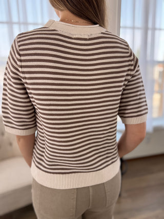 A woman viewed from behind wearing a brown and cream horizontally striped short-sleeve knit top, paired with beige pants, in a brightly lit indoor setting.
