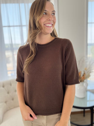 A woman wearing a short-sleeve brown knit sweater and beige pants, standing in a bright, modern room with large windows and a tufted sofa in the background.