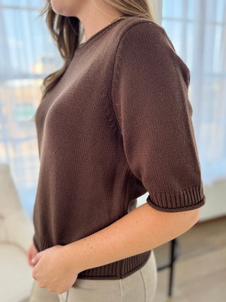 A woman wearing a short-sleeve brown knit sweater with ribbed cuffs and hem, paired with light beige pants, shown in a softly lit indoor setting.
