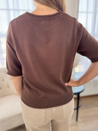 A woman viewed from the back wearing a brown short-sleeve knit sweater and light beige pants in a bright indoor room.