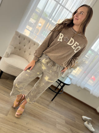 A woman modeling a brown long-sleeve sweatshirt with 'RODEO' printed in beige across the chest, paired with distressed gray pants and beige platform sandals, in a bright room with a tufted sofa and large windows.