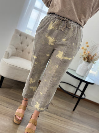 A person wearing loose, cropped drawstring pants in a taupe-gray color with a distressed beige splatter pattern, paired with brown platform sandals in a bright living room setting.