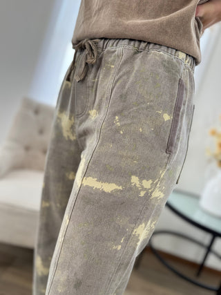 Close-up of a person wearing faded taupe drawstring sweatpants with a light beige tie-dye or bleach-splatter pattern, paired with a matching taupe top in a bright indoor setting.