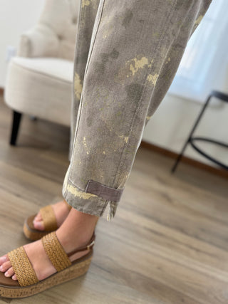 Close-up of a person's legs wearing cuffed beige and brown patterned pants with woven tan wedge sandals, standing on a light wood floor in a bright indoor room.