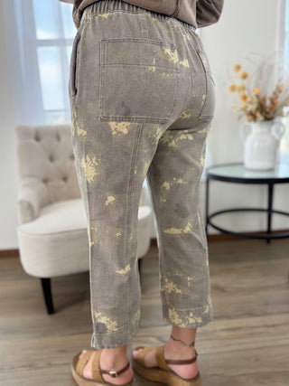 A person shown from the back wearing cropped gray pants with a distressed gold-yellow bleach-splatter pattern, paired with brown sandals in a bright indoor room.