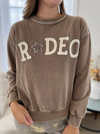 A person wearing a brown washed sweatshirt with 'RODEO' and a star motif in cream and leopard print on the front, paired with distressed gray jeans.
