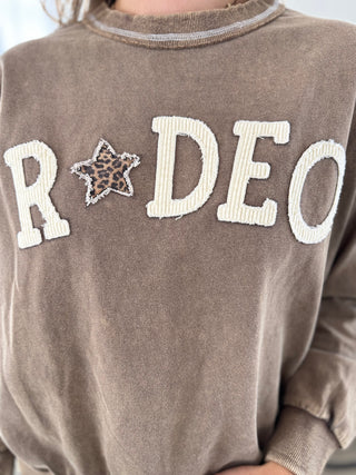 A close-up of a person wearing a brown long-sleeve sweatshirt with 'RODEO' embroidered across the chest in cream ribbed letters, with a leopard-print star between the letters.