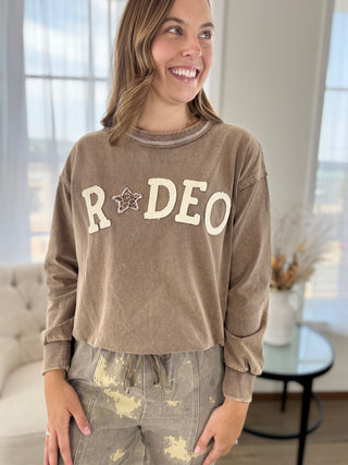 A woman wearing a brown cropped long-sleeve sweatshirt with 'RODEO' and a star graphic on the front, paired with distressed gray pants, standing in a brightly lit room.