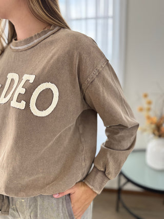 A person wearing a brown long-sleeve sweatshirt with large cream-colored 'VIDEO' lettering on the front, shown in a softly lit indoor setting.