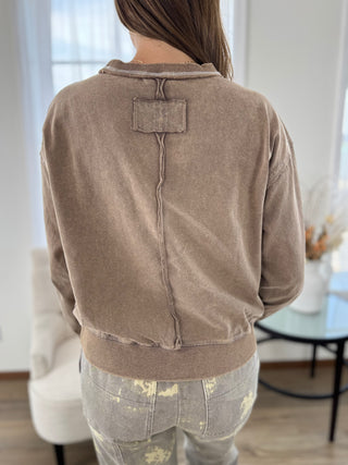 A person shown from the back wearing a taupe-brown cropped sweatshirt with a unique stitched back detail and light gray paint-splattered jeans in a bright indoor setting.