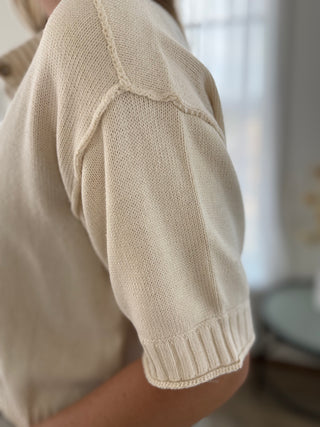 Close-up of a person wearing a cream-colored short-sleeve knit top, showing the ribbed sleeve cuff and shoulder seam in soft natural light.