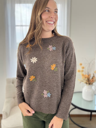A woman wearing a brown knit sweater with colorful embroidered flowers, paired with olive green pants, standing in a bright, modern living room.