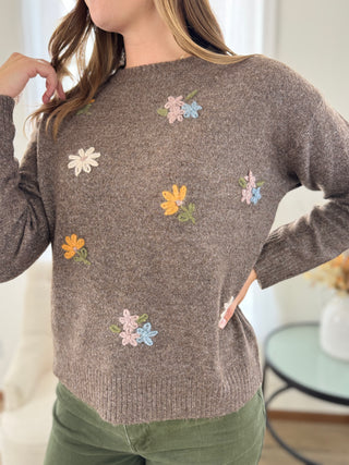 A woman wearing a taupe-brown knit sweater with colorful embroidered flowers, paired with olive green pants, in a brightly lit indoor setting.