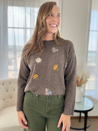 A woman wearing a brown knit sweater with colorful embroidered flowers, paired with olive green pants, standing in a bright, modern living room.
