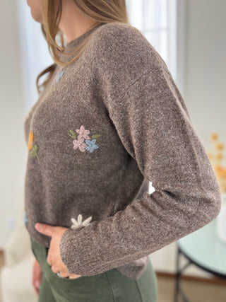 A woman wearing a heathered taupe-brown knit sweater with colorful embroidered flowers on the chest, paired with olive green pants, shown in a bright indoor setting.